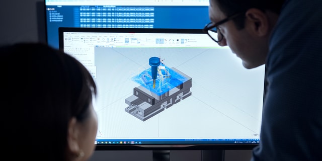 A woman and a man looking at a computer screen with an image of a toolpath for a milling operation which is generated using CAM software