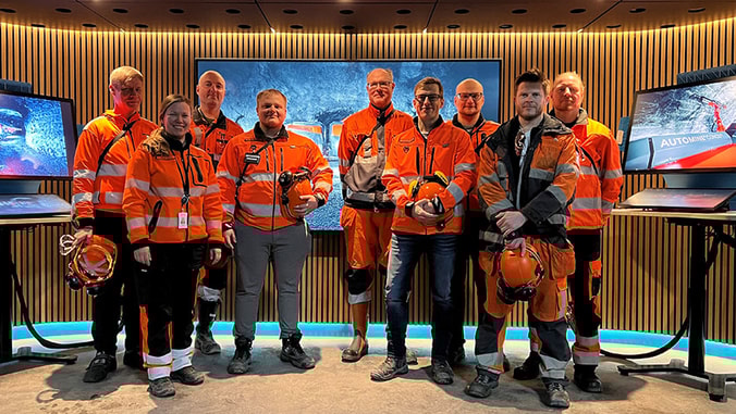 Nine people wearing personal protective equipment for mining operations standing next to each other with three large monitors behind them.
