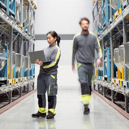 A woman holding a laptop and a man walking between two warehouse shelfs