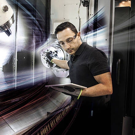A man standing next to a CNC machine looking on a tabket that he holds in one of his hands
